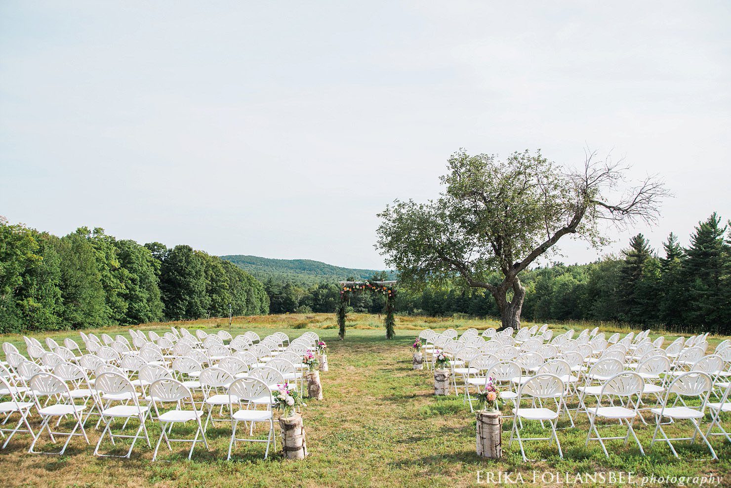 Wedding ceremony in an open field with a single apple tree, white birch arbor, and views of rolling hills | Henniker, NH